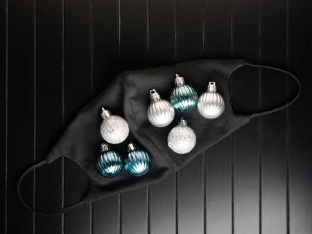 Black face mask decorated with christmas ornaments on black wooden background. Top view