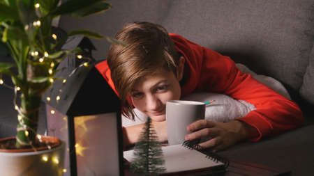 Cute boy with cup lying on the sofa and drawing picture. Evening time christmas lights and decorationsの写真素材