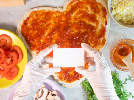 Hands holding empty business card on hear shape pizza dough with tomato sauce background. Ingredients for cooking pizza with dough in heart shapeの写真素材