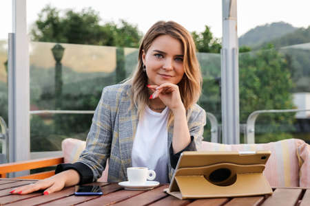 Charming young business lady sitting at the table with tablet and cup of coffee in outdoor cafe. Looking at cameraの写真素材