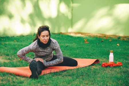 Young woman stretching and warming up on the mat, touching the foot, on green grass outdoors