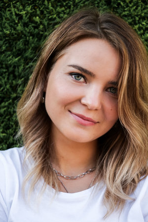 Summer outdoor portrait of young woman in white shirt with natural makeup looking at camera. Standing by green hedgeの写真素材