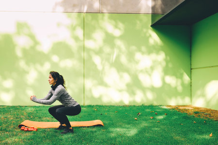 Sporty young woman doing squat exercise outdoors on the grass near green wall. Wearing warm sport clothesの写真素材