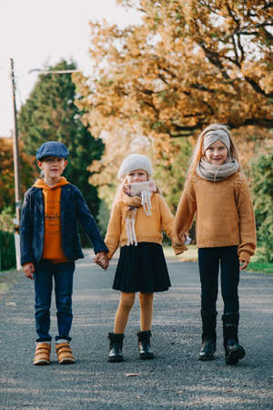 Adorable happy kids holding hands standing on the road in autumn day. Brother and two sisters family portrait outdoorsの写真素材