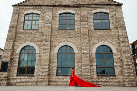 Young charming woman in long red dress and red shoes walking gorgeously in the city by brick buildingの写真素材