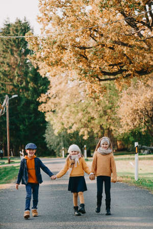 Adorable happy kids holding hands standing on the road in autumn day. Brother and two sisters family portrait outdoorsの写真素材