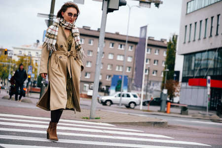 Young woman wearing beige coat, scarf and sunglasses crossing the road