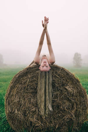 Young woman in beige dress lying on haystacks in mist field at twilight time looking upの写真素材