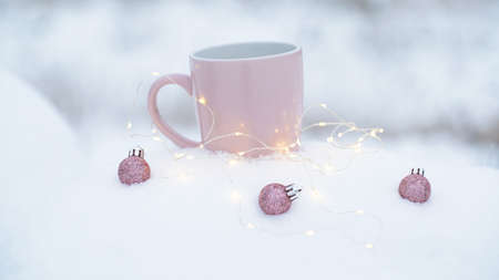 Snow is falling. Closeup of cup of hot drink with christmas balls and lights on snow.の写真素材
