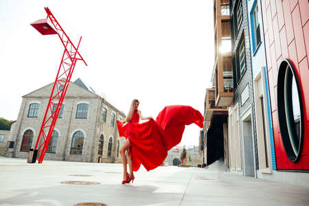 Young charming woman in long flying red dress and red shoes posing on the modern new streetの写真素材