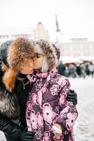 Daughter and mother in warm clothes kissing outdoors at Christmas market. Happy holidays and merry Christmasの写真素材