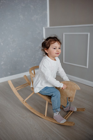 Cute emotional two and half years old child girl swinging on a wooden horse indoors. Happy childhoodの写真素材
