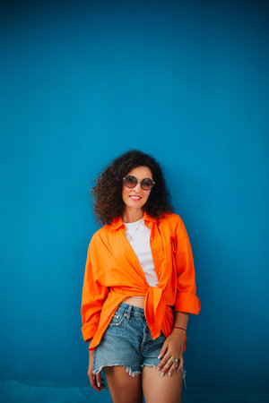 Bright and colorful. Stunning curly haired woman in shorts and orange shirt posing against violet wall.の写真素材