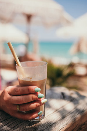Woman hand holds Ice coffee latte on the beach. Summer iced coffee latte in tall glass with straw by the sea in beach bar coffeeの写真素材