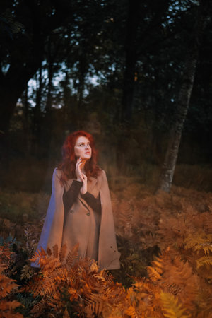 Red-haired woman in beige coat in autumn forest. Beautiful dreamy lady standing in ferns outdoors and looking thoughtfully aside. Lens flare effectの写真素材