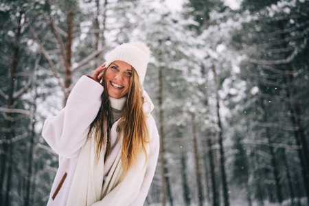 Active beautiful young woman in warm white sheepskin coat, hat and mittens and flying scarf enjoying snowy winter forest, looking at cameraの写真素材