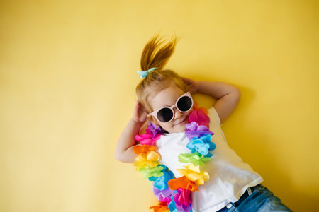 Adorable little girl in white t-shirt wearing summer sunglasses lying on yellow background, studio portrait. Summer vacationsの写真素材