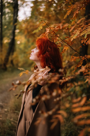 Closeup side profile portrait of a beautiful autumn woman with dyed red hair and pierced nose standing near colorful autumn leaves. Pretty woman revealing individuality.の写真素材