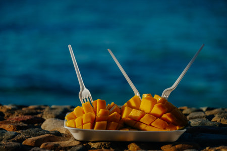 Fresh sliced mango fruit with plastic forks on the shore by the sea. Summer vacationの写真素材