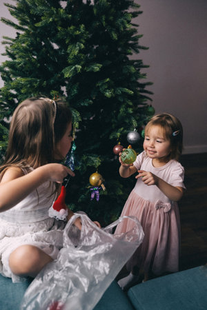 Sisters in beautiful dresses decorating artificial Christmas tree with balls and toys. Cozy Christmas homeの写真素材