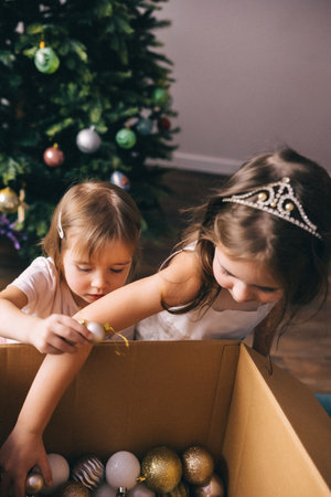 Sisters in beautiful dresses decorating artificial Christmas tree with balls. Cozy Christmas homeの写真素材