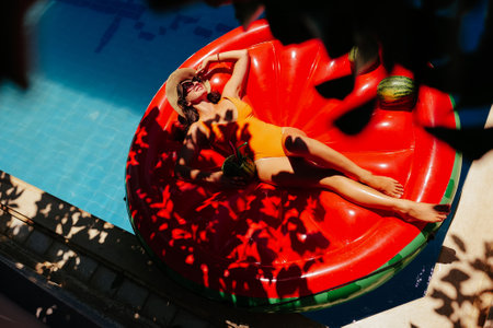 High angle paparazzi view from the tree branches on pretty woman in orange swimsuit and sunglasses lying on inflatable watermelon island with small ripe watermelon fruits on it in swimming poolの写真素材