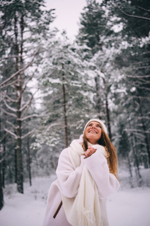 Active beautiful young woman in warm white woolen jacket, hat and mittens and flying scarf enjoying snowy winter forest, looking aheadの写真素材
