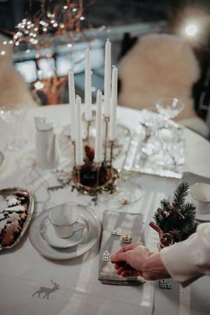 Woman hand setting up table for Christmas dinner. Table is decorated with white tablecloth, cups, glasses, candlestick, table-napkin and small wooden decorative housesの写真素材