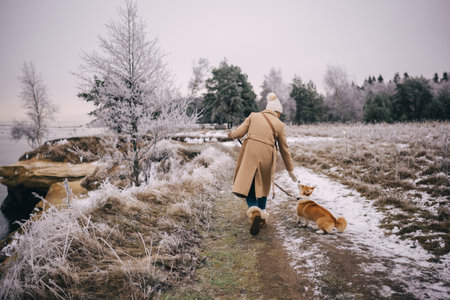 Happy attractive young woman wearing beige coat, white hat and mittens walking with welsh corgi pembroke puppy, outdoors near winter sea. Backside viewの写真素材