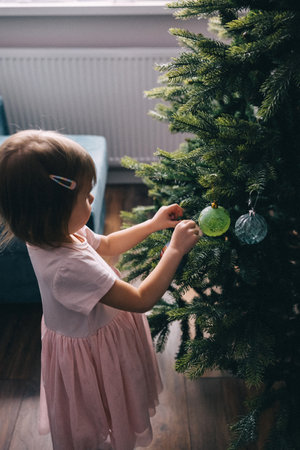 Adorable two years old toddler girl in coral pink dress decorating Christmas tree with balls. Cozy Christmas homeの写真素材