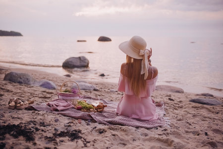 Young woman in straw hat and pink dress sit on plaid have picnic on sea sunrise sand beach. Mind and soul healing power of the sea.の写真素材