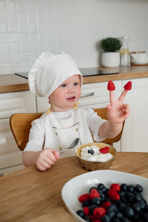 Adorable 3 years old boy in chefs hat and apron puts raspberries on fingers. Kids cooking at kitchenの写真素材