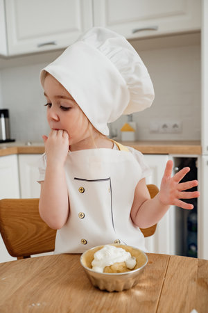 Adorable girl in chef hat and aprons preparing cake at the kitchen. Child licking fingers. Delicious sweet food.の写真素材