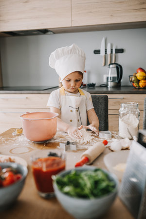 Adorable girl in chefs hats and aprons playing with the flour in the kitchen, preparing pizza. Kids hobbiesの写真素材