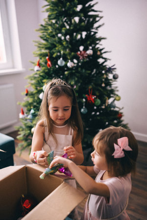 Sisters in beautiful dresses decorating artificial Christmas tree with baubles and toys. Cozy...