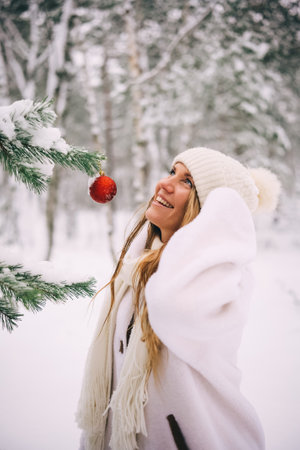 Young happy woman in white sheepskin coat, hat and scarf standing near fir tree with red ball in snowy winter forest looking ahead dreamlike. Cozy and happy Christmas and New Year holidaysの写真素材