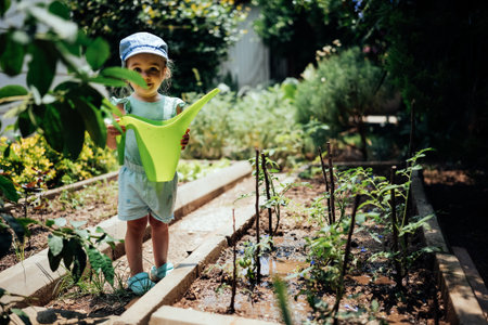 Adorable girl in summer clothes holding watering can standing between garden beds looking at camera....