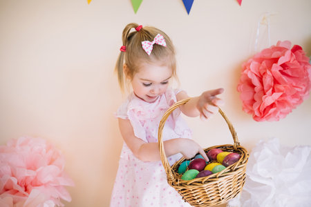 Cute happy 3 years old girl wearing pink dress holding basket with painted Easter eggs in bright...