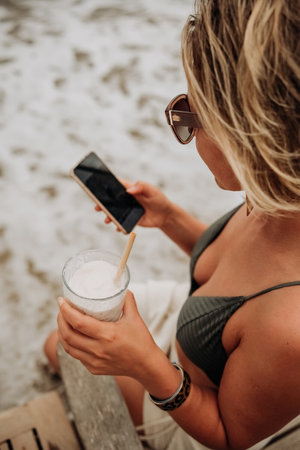Young woman in bikini on the beach using mobile phone and drinking cocktailの写真素材