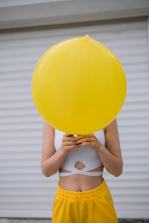 Cheerful young woman wearing yellow pants hiding behind yellow balloon standing on the street