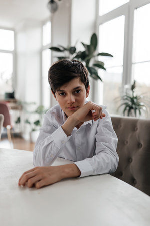 A young boy rests his chin on his hand while sitting at a table in a bright, plant-filled room, deep in thought.の写真素材