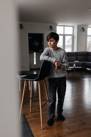 A young boy leans against a tall chair, looking thoughtful in a spacious, well-lit living room with wooden floors.の写真素材