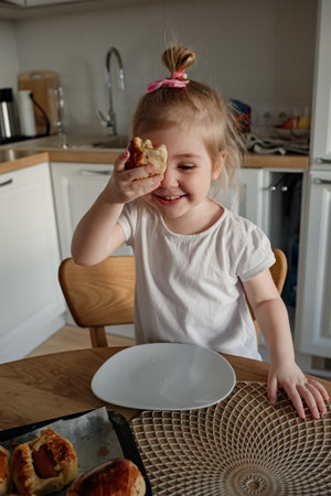 A cheerful young girl smiles as she holds a delicious pastry in one hand, sitting at a bright kitchen table, ready to enjoy her treat.の写真素材