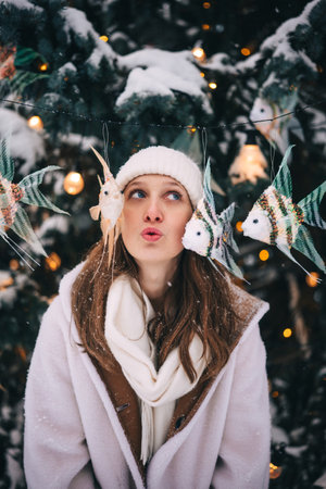 Funny young woman stands in the snow, wearing warm clothing, with colorful decorations hanging around her.の写真素材