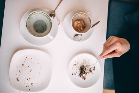 A hand reaches for leftover dessert crumbs while coffee cups rest on a table in a cafe.の写真素材