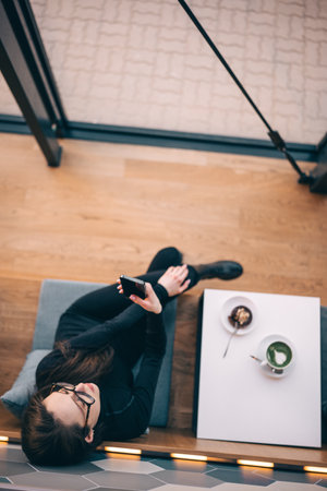 Adorable young woman wearing glasses drinking coffee or tea and using smartphone at cozy cafe....
