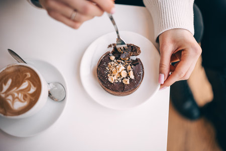 A hand holds a fork, taking a bite of a chocolate dessert beside a cup of coffee in a cafe.