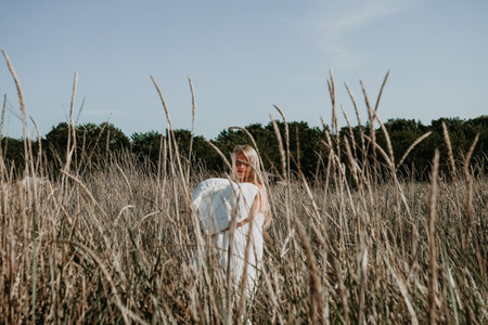 Young blonde woman walks through a field of tall grass wrapped in white blanket in the morning....