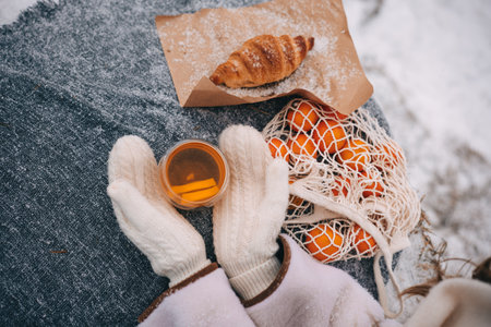 Hands hold a warm drink alongside a croissant and a bag of oranges on a winter blanket.の写真素材