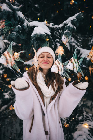 Funny young woman stands in the snow, wearing warm clothing, with colorful decorations hanging around her.の写真素材